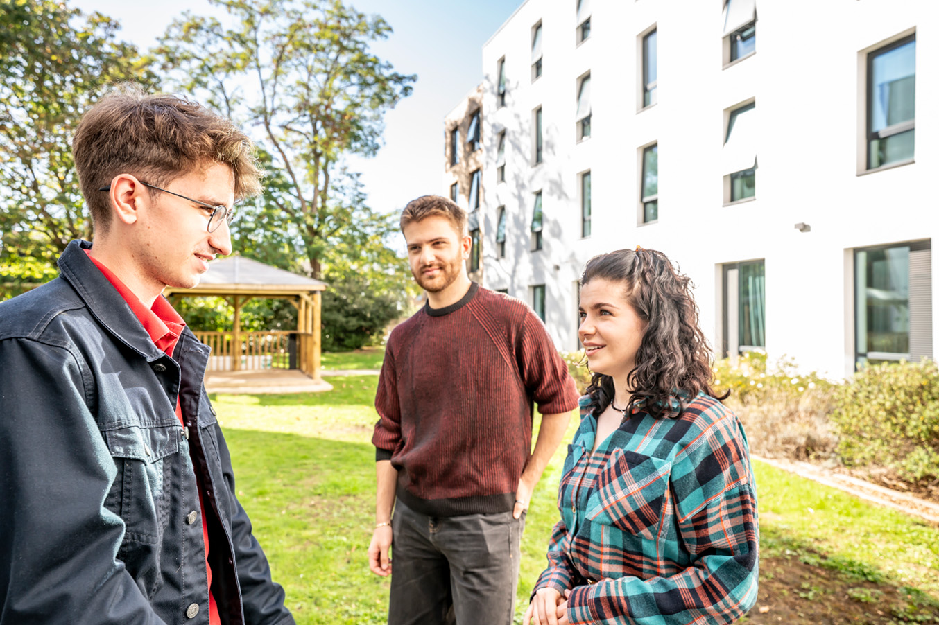 Three students, wearing casual clothing, chatting outside a white building, in a green area.